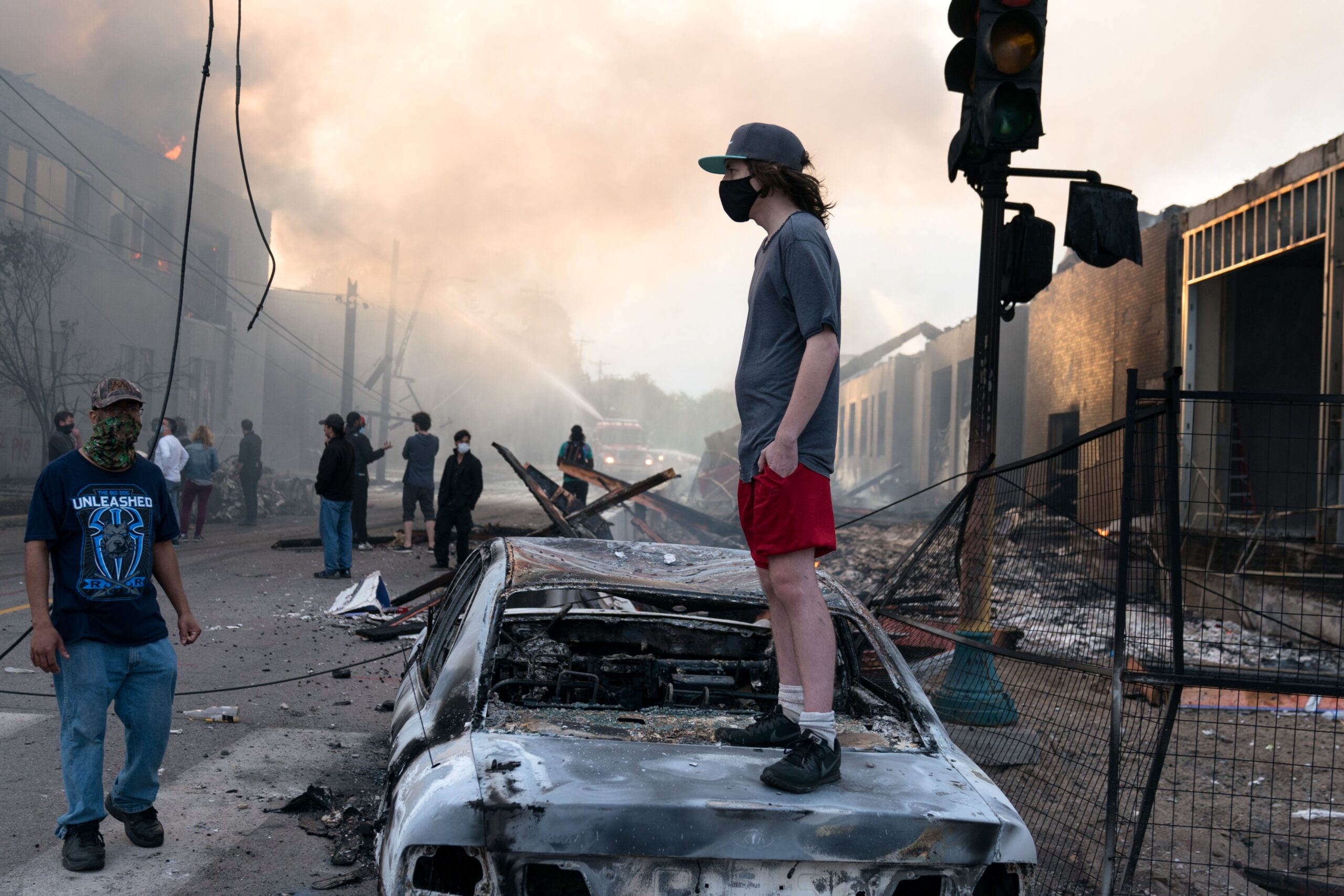 A young man stands on a burned-out car amidst a scene of destruction, smoke, and a crowd in the background during a crisis event.