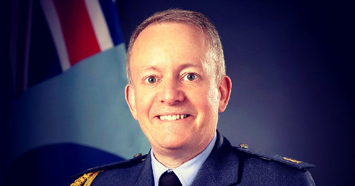 Portrait of a smiling military officer in uniform against a backdrop featuring national flags, conveying professionalism and leadership.