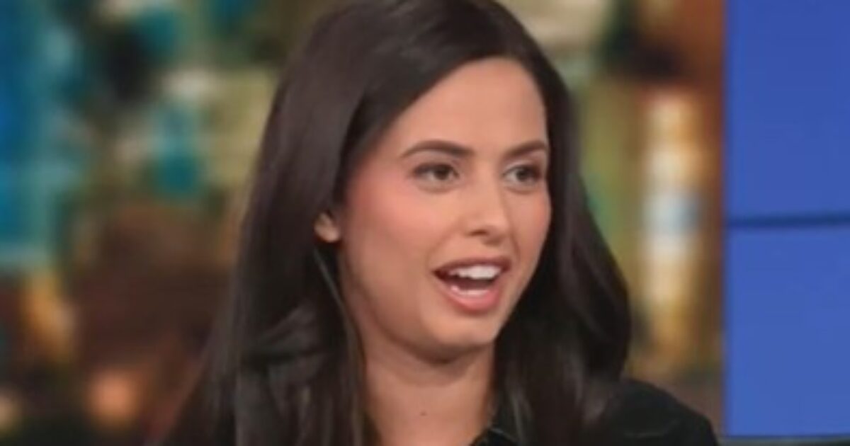 Woman with long dark hair smiling and speaking during a television interview, set against a blurred urban background.
