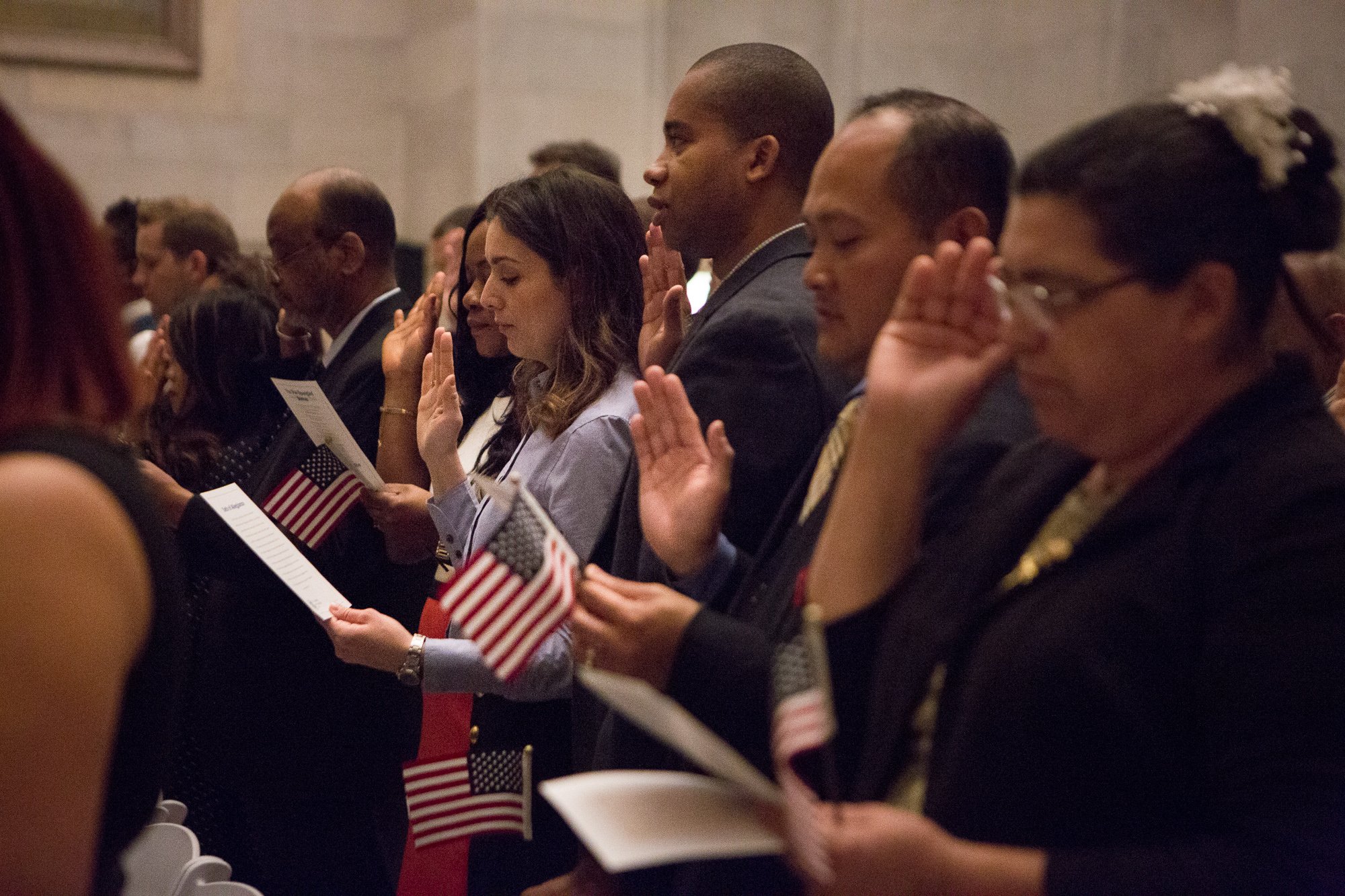 Diverse group of new citizens taking the Oath of Allegiance while holding small American flags during a naturalization ceremony.