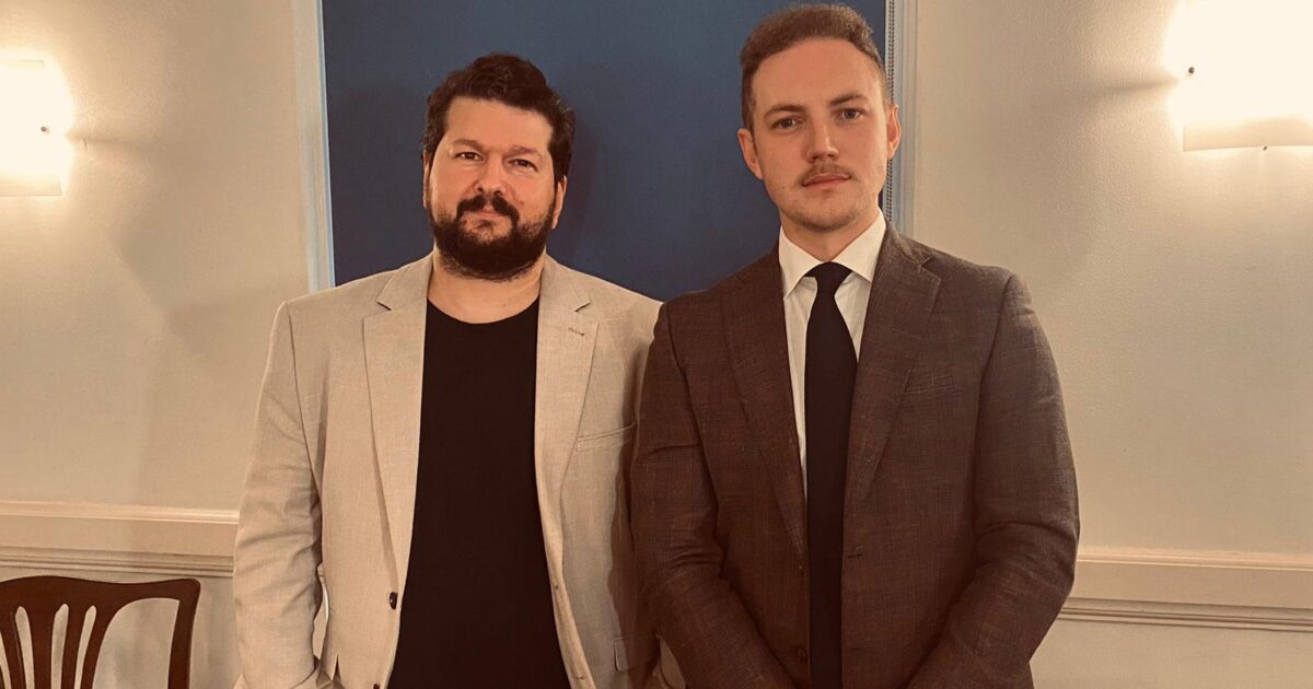 Two men pose for a photo in formal attire, one in a light blazer and the other in a dark suit, against a softly lit background.