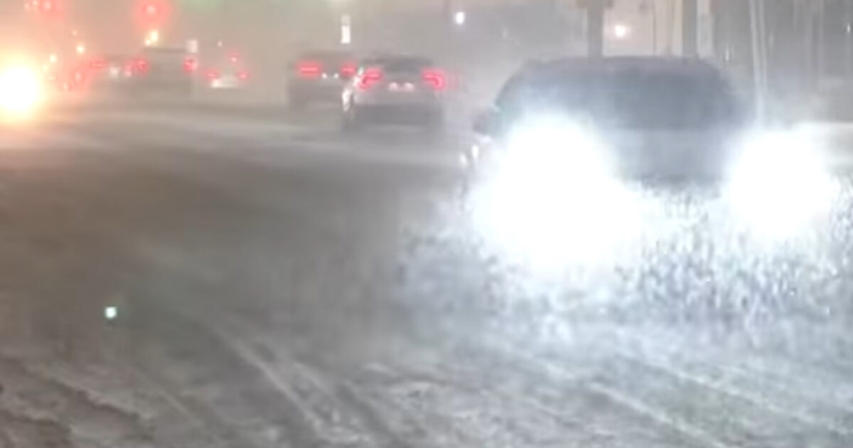 Heavy snowfall creates low visibility conditions as vehicles navigate a busy street at night.