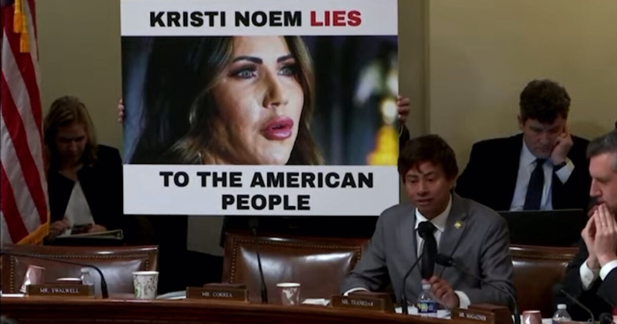 A congressional hearing featuring a sign accusing Kristi Noem of lying to the American people, with lawmakers engaged in discussion.