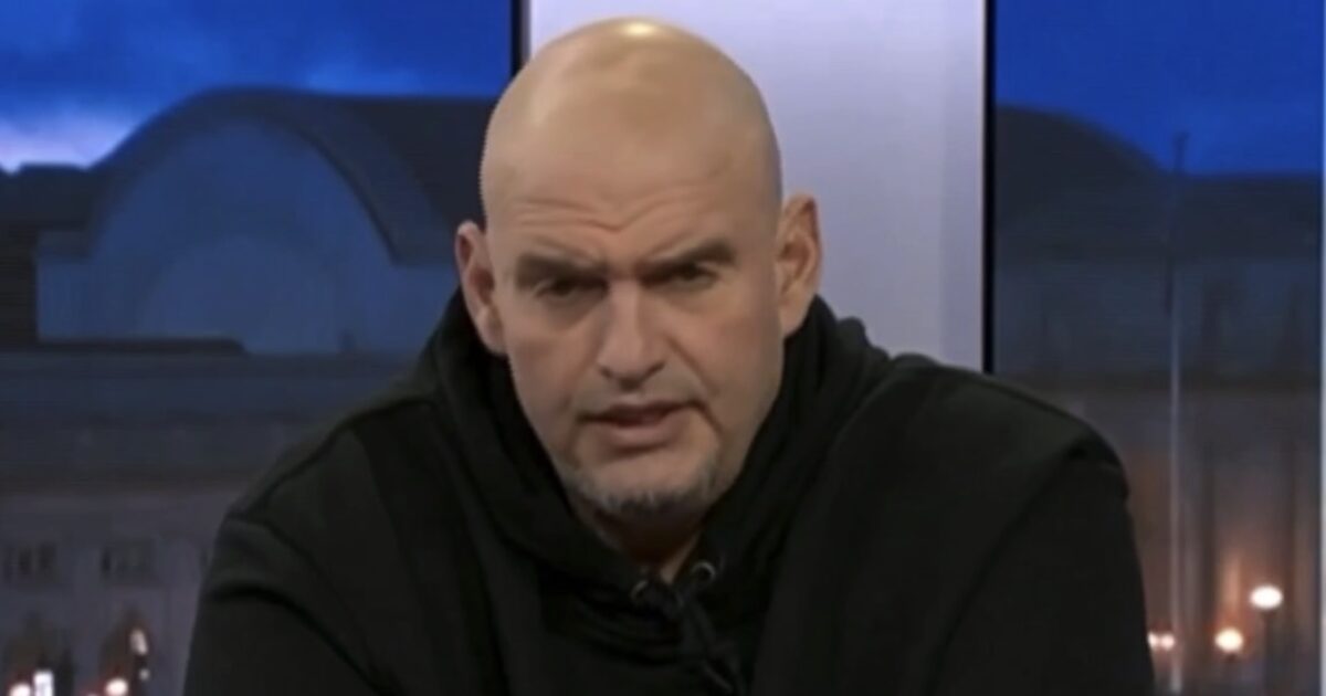 Bald man in a black hoodie speaking intently during a television interview, with a blurred background of a cityscape at dusk.