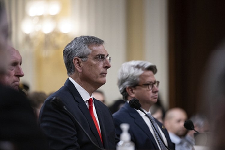 Two men in formal attire sit attentively during a congressional hearing, with microphones in front of them and an audience visible in the background.