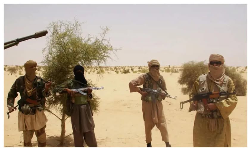 Armed individuals in desert terrain, displaying military attire and weapons, standing near sparse vegetation.