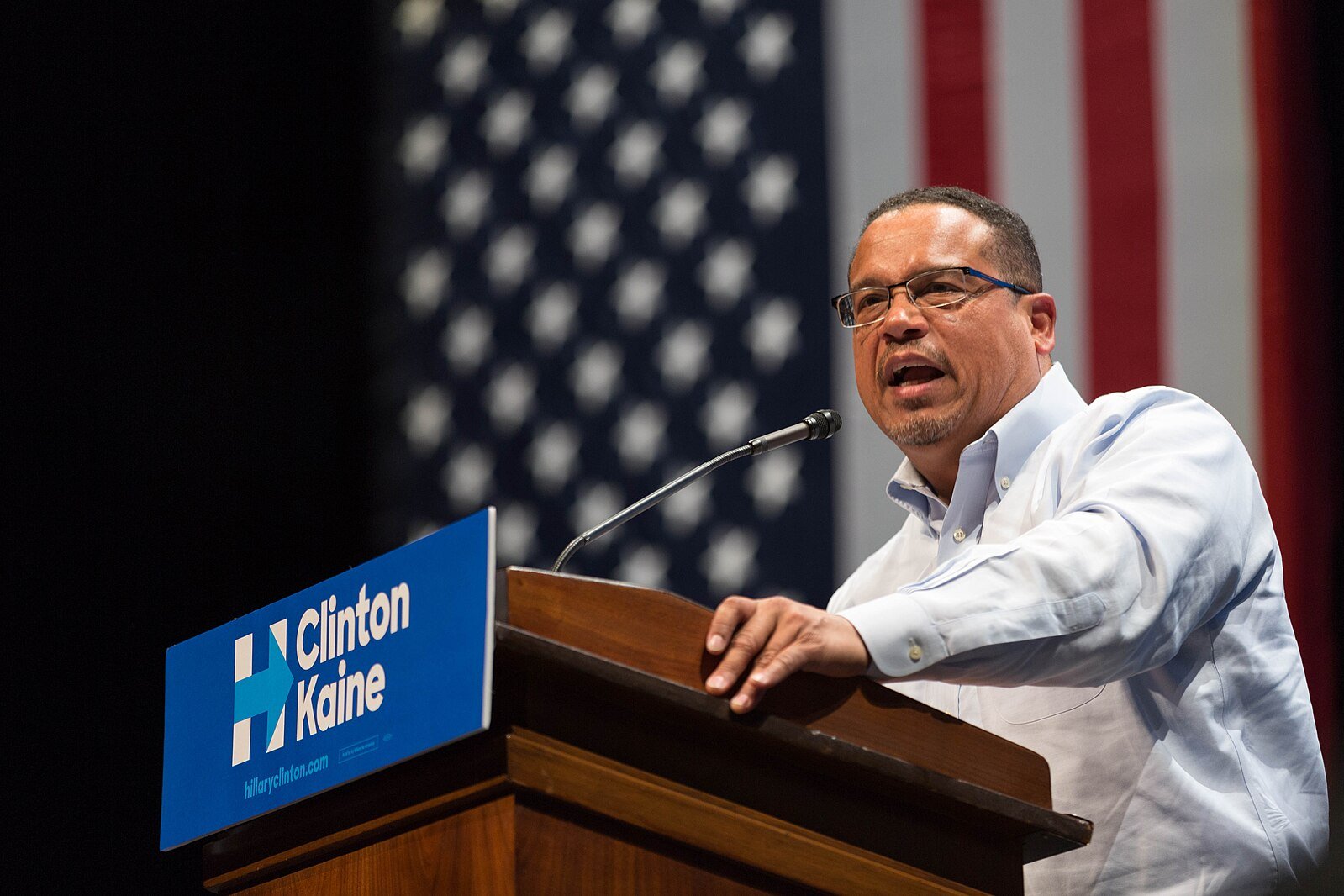 Political speaker addressing an audience at a rally with a Clinton-Kaine sign and American flag backdrop.
