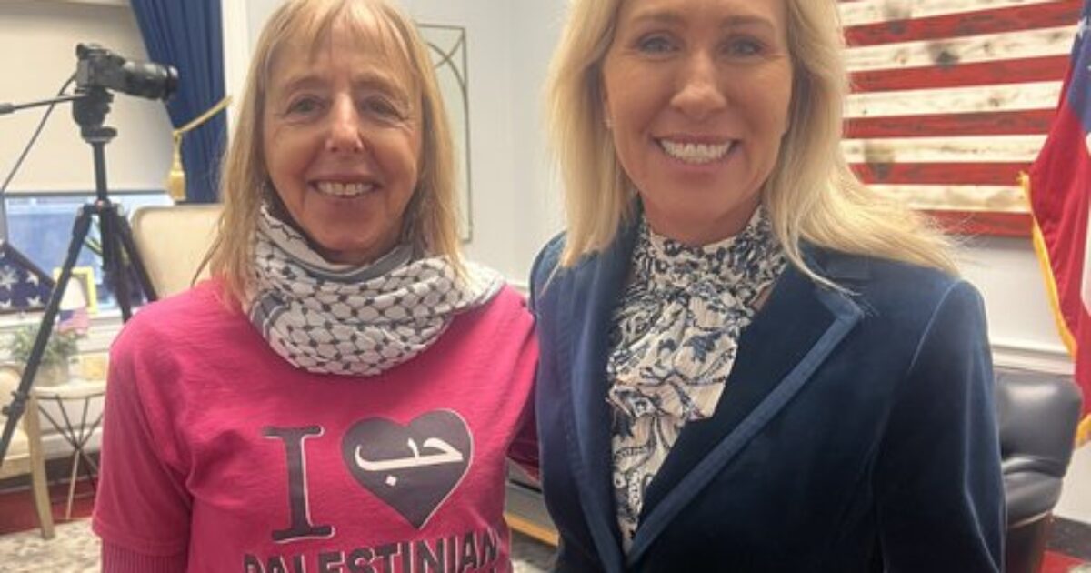 Two women pose for a photo in an office setting, one wearing a pink shirt with a pro-Palestinian message and the other in formal attire.
