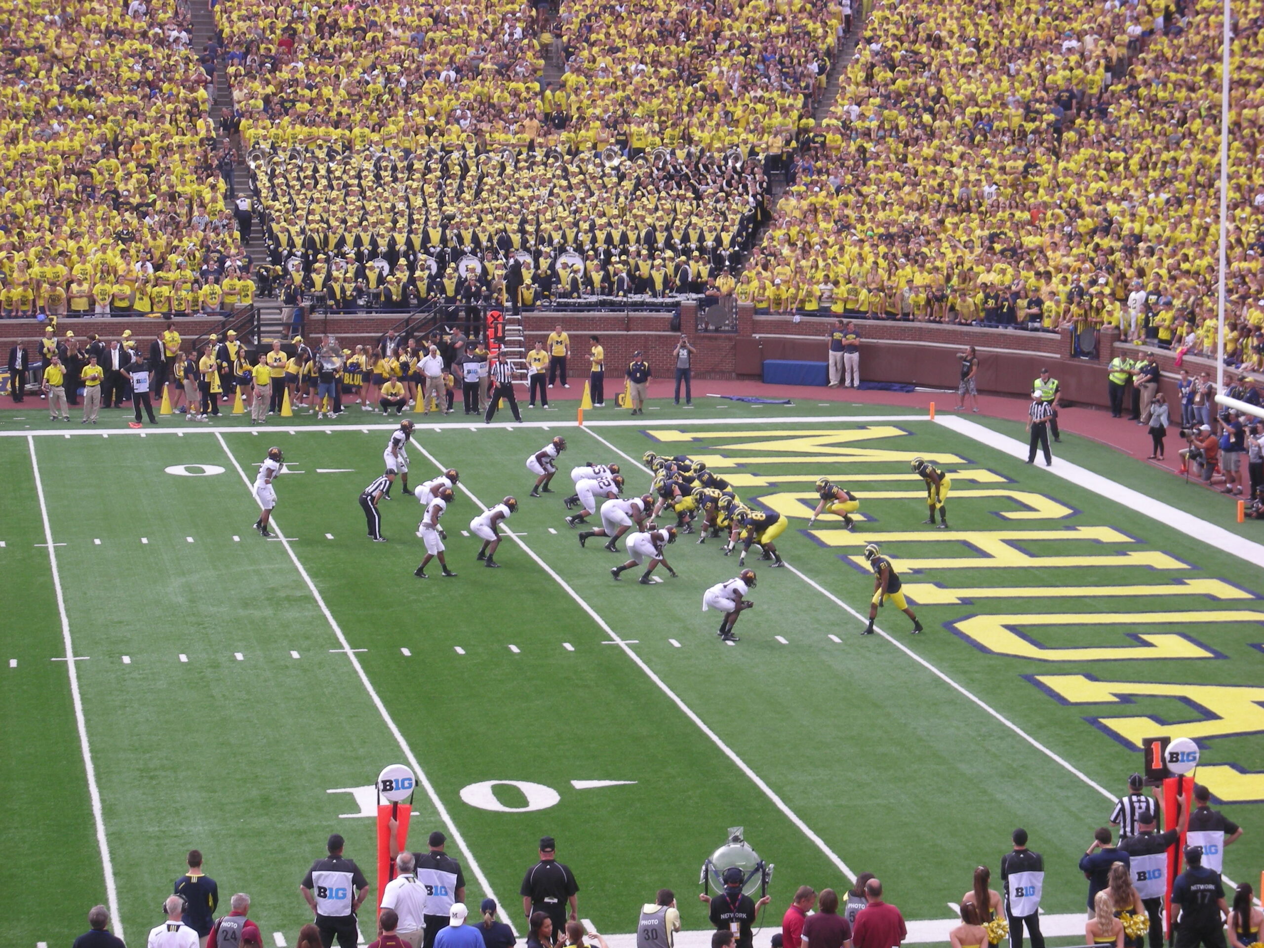 A college football game at Michigan Stadium featuring players in action with a vibrant crowd dressed in maize and blue.