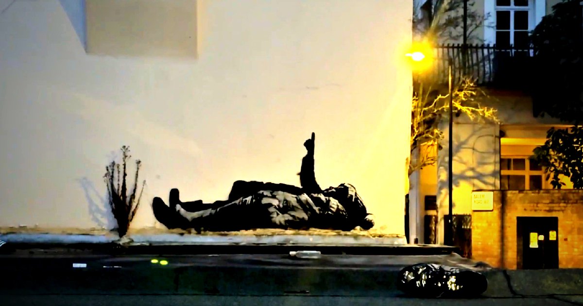 Street art depicting a figure lying on the ground, illuminated by nearby streetlights, against a white wall with urban surroundings.