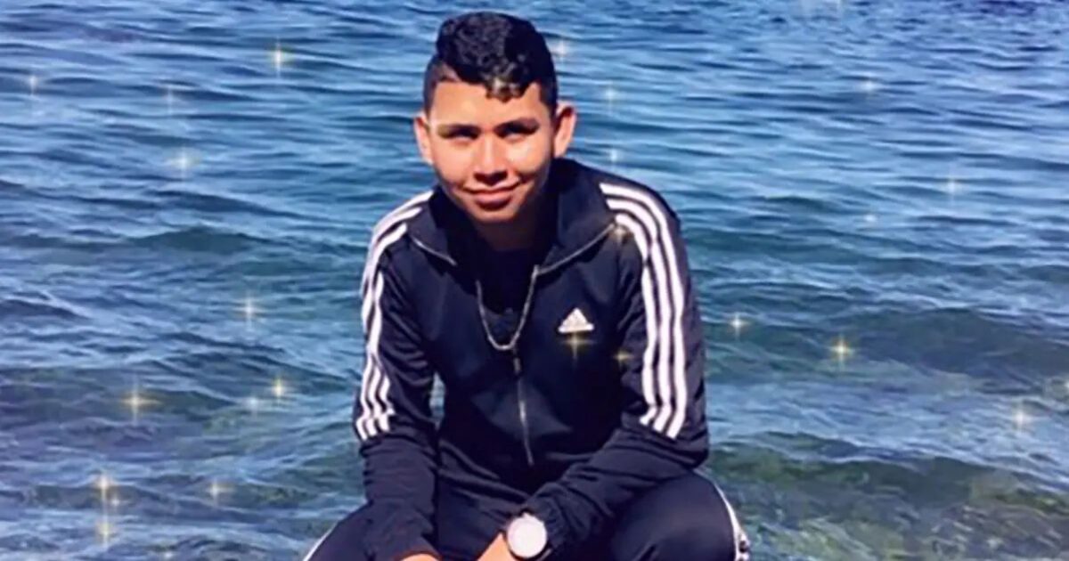 Young man in a black tracksuit crouching by the water, smiling with a scenic background of sparkling waves.