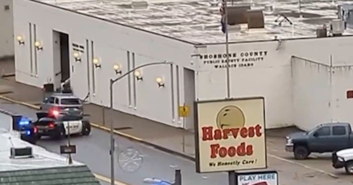 Police cars parked outside the Hoshone County Public Safety Facility in Wallace, Idaho, with a Harvest Foods store visible in the foreground.