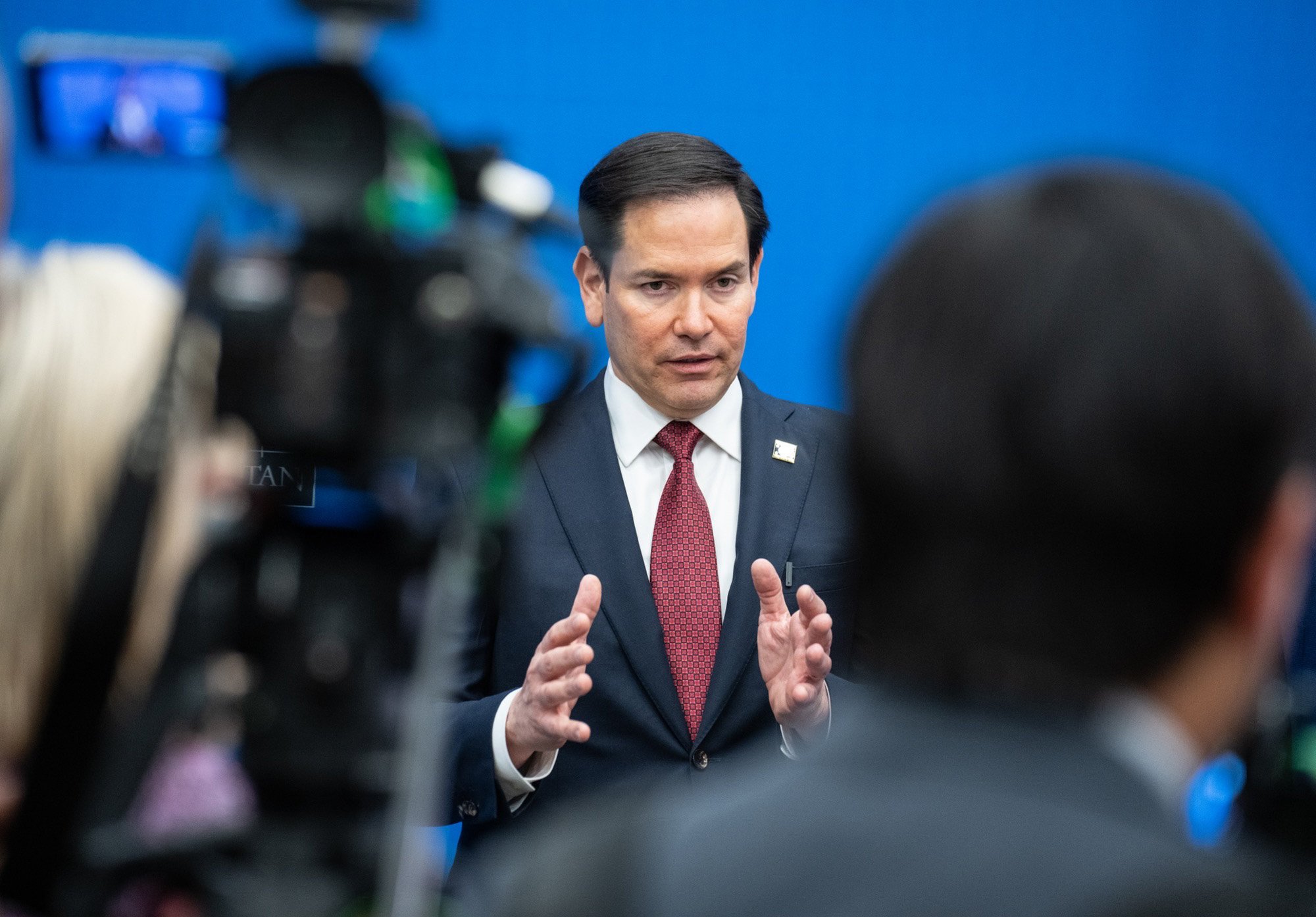 U.S. Senator Marco Rubio speaks during a press event, gesturing with his hands against a blue backdrop and surrounded by cameras.