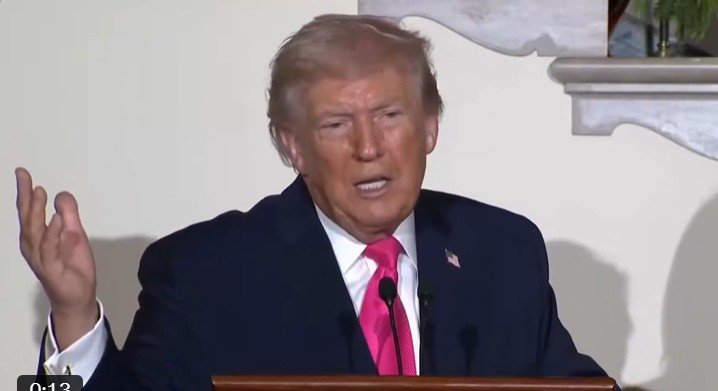 Donald Trump delivering a speech at a podium, wearing a dark suit and pink tie, gesturing with his hand.