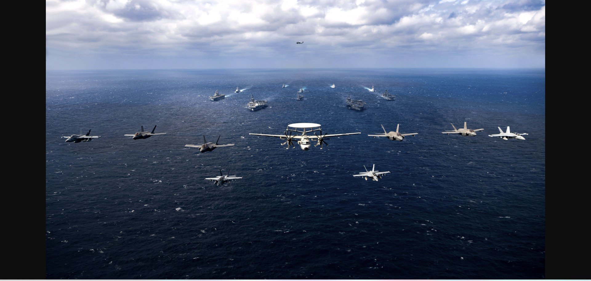 Military aircraft flying over a naval fleet in the ocean under a cloudy sky, showcasing aerial operations and maritime power.