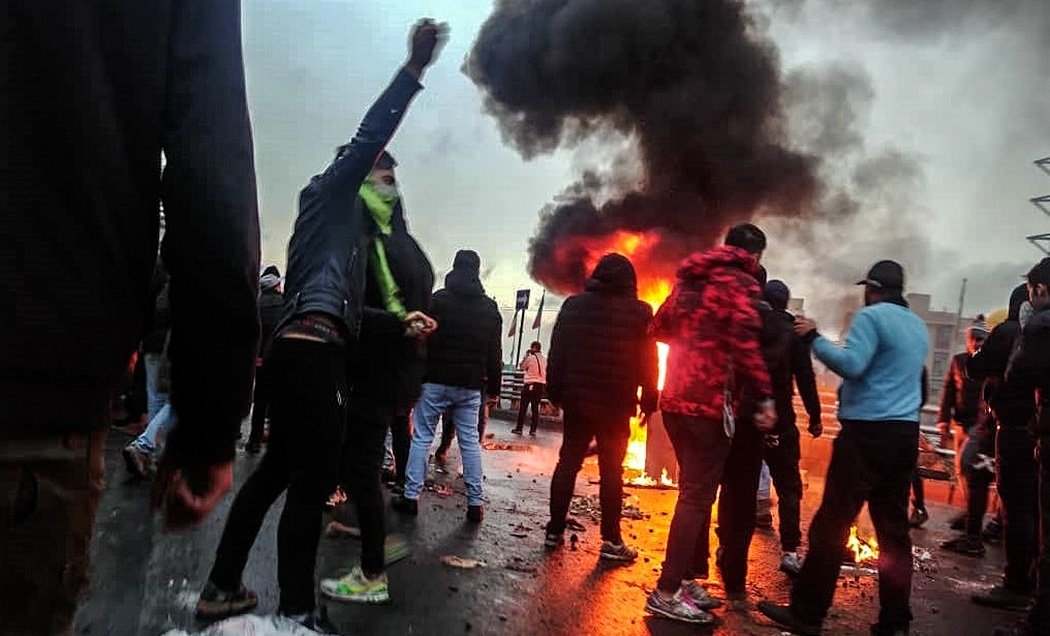 Protesters gather around a fire, raising their fists in defiance against a backdrop of smoke and tension during a demonstration.