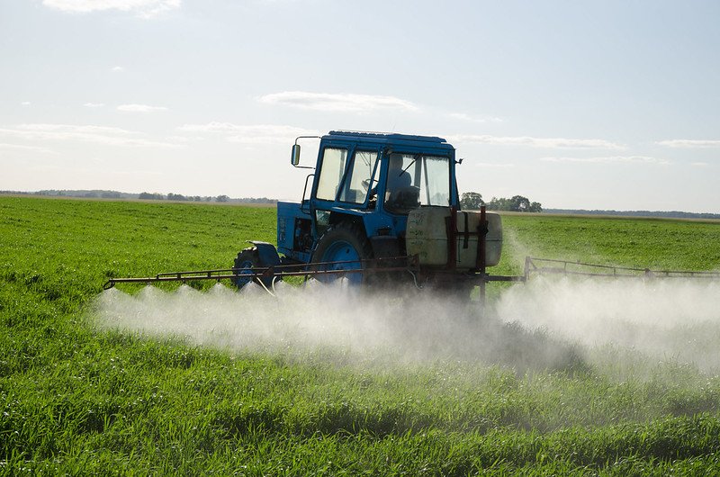Blue tractor spraying pesticides on a lush green field under a clear sky.