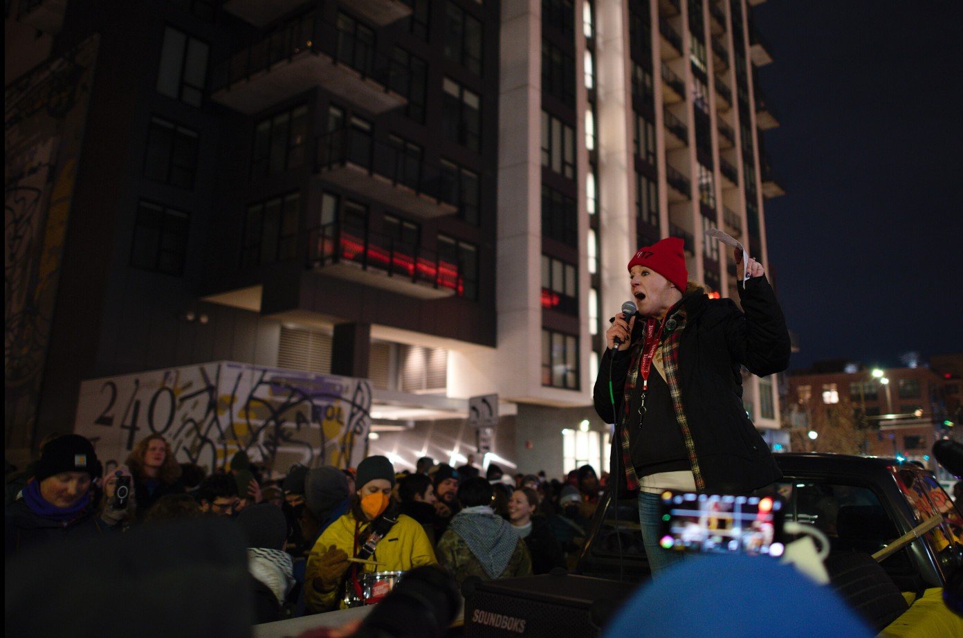 A speaker passionately addresses a crowd during a nighttime protest, with urban buildings and graffiti visible in the background.