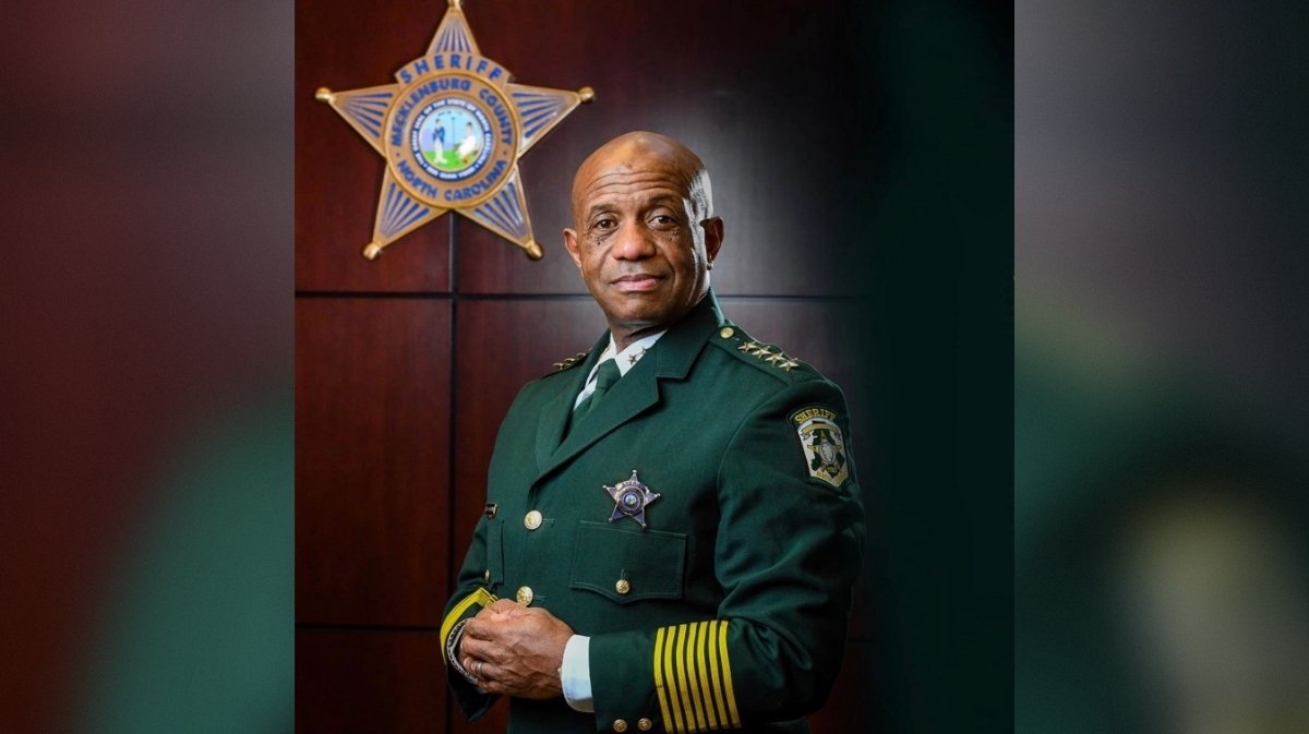 Portrait of a sheriff in a green uniform, standing confidently with a badge, in front of a wooden backdrop featuring a sheriff's star emblem.