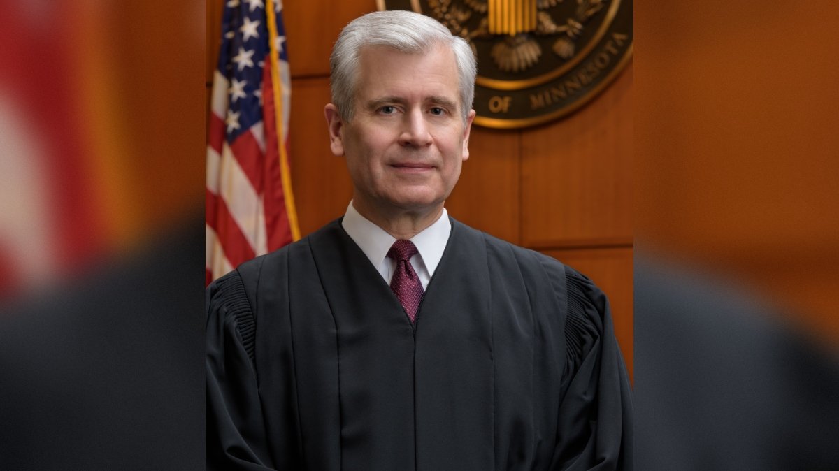 Portrait of a judge in a black robe standing in a courtroom with American flags and a seal of Minnesota in the background.