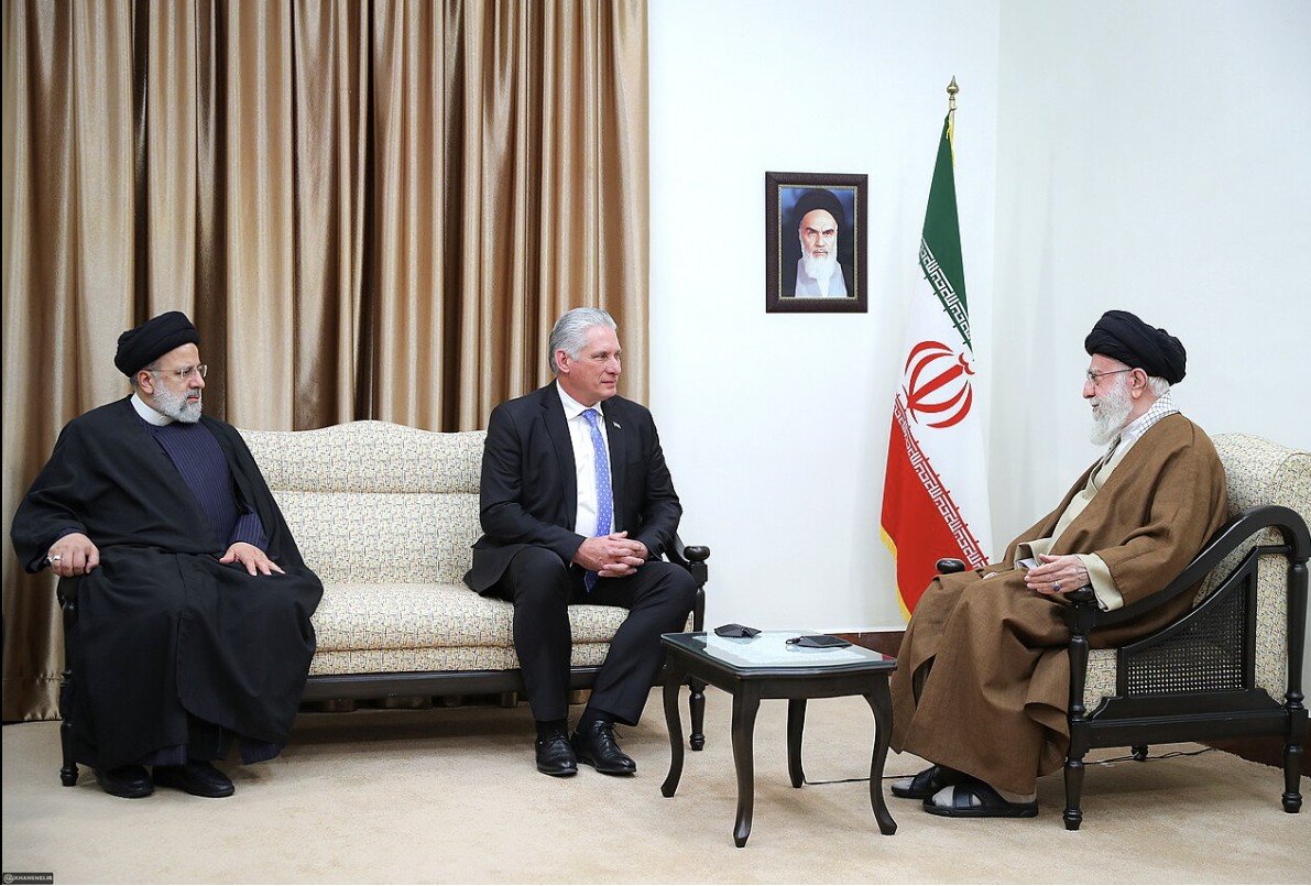 Meeting between Iranian leaders Ebrahim Raisi and Ali Khamenei with a foreign dignitary, discussing diplomatic relations, with the Iranian flag and a portrait in the background.