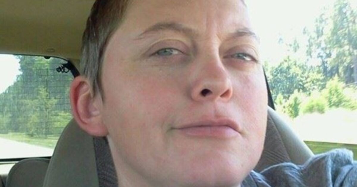 Close-up of a woman with short hair, sitting in a car, looking directly at the camera with a neutral expression, surrounded by greenery outside.