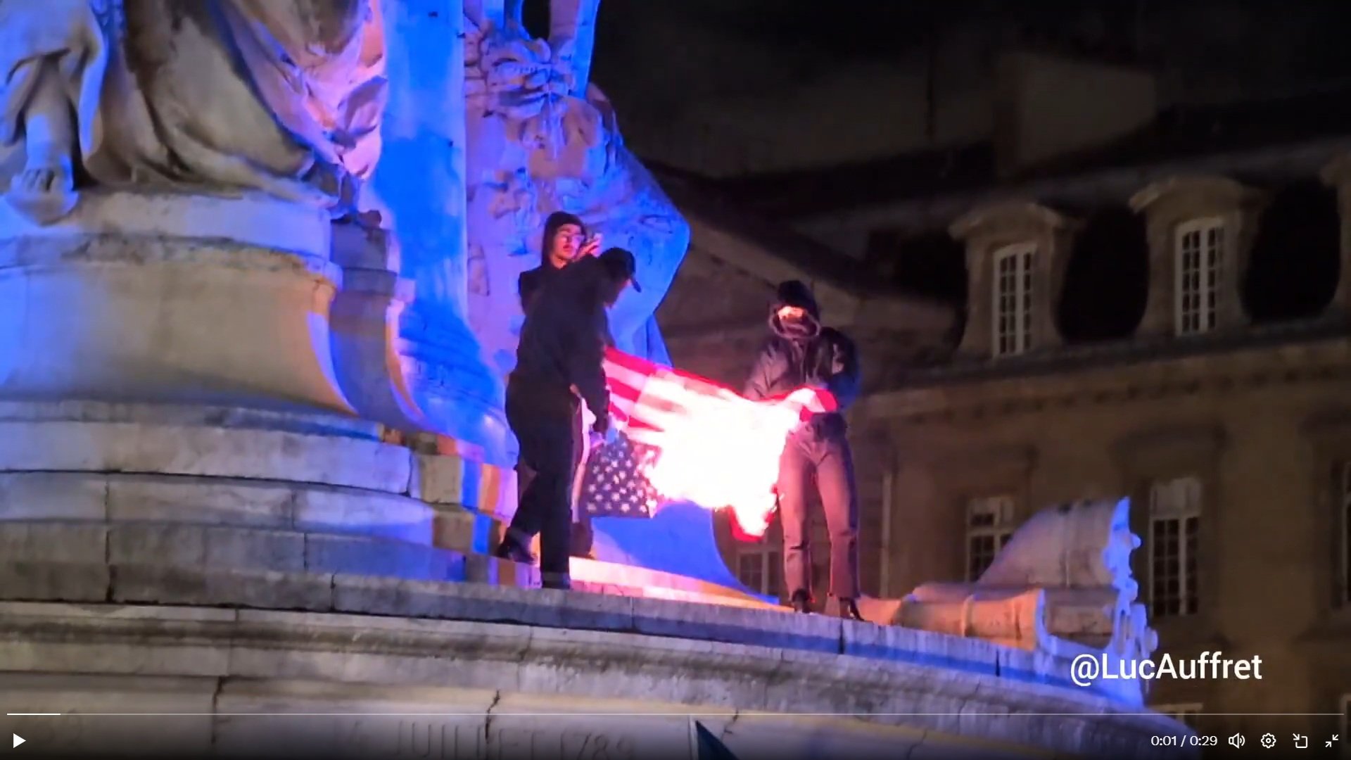 Protesters burn an American flag on a monument at night, illuminated by blue lights, highlighting a controversial political demonstration.