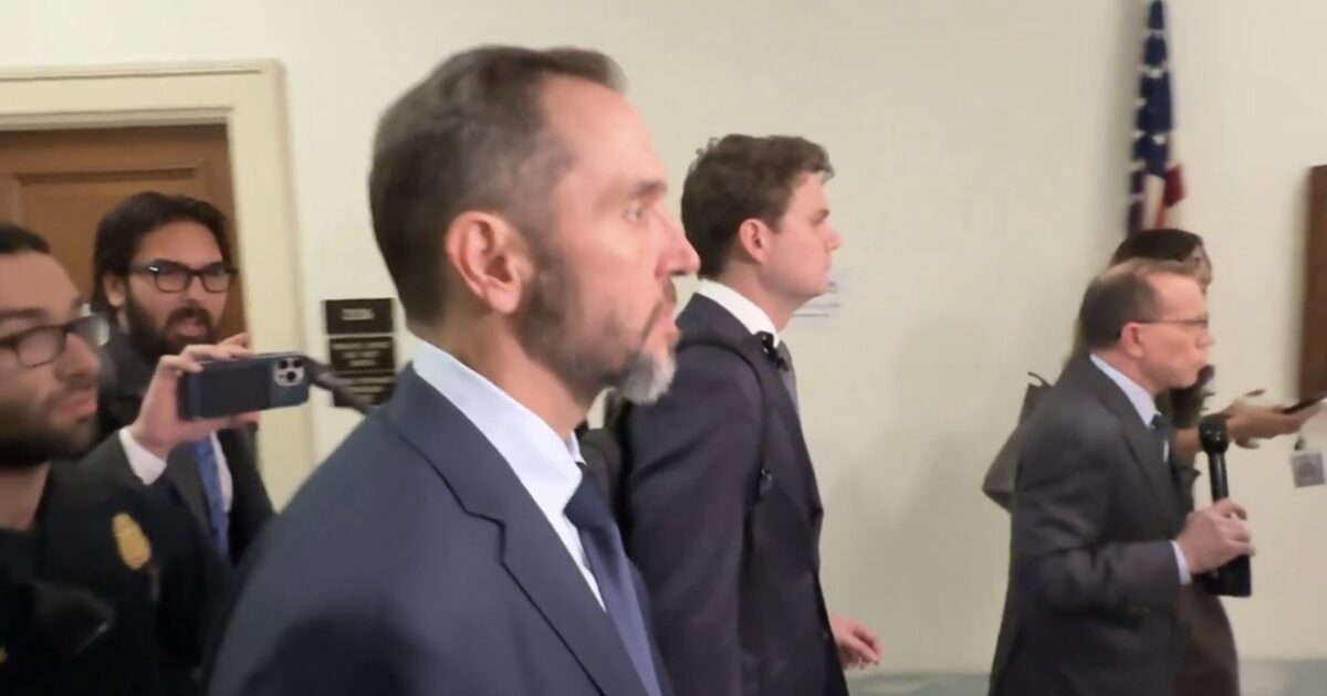 Group of individuals, including a man in a suit, walking through a hallway while being filmed by onlookers during a public event.