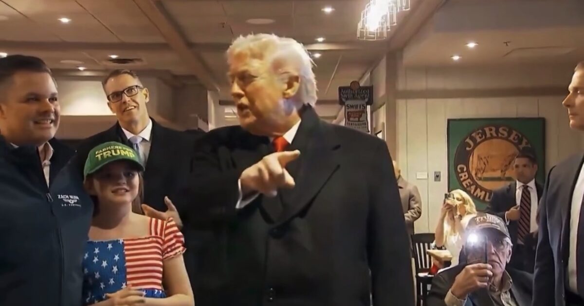Donald Trump engages with supporters in a casual setting, surrounded by attendees, including a young girl in an American flag outfit and a man taking photos.