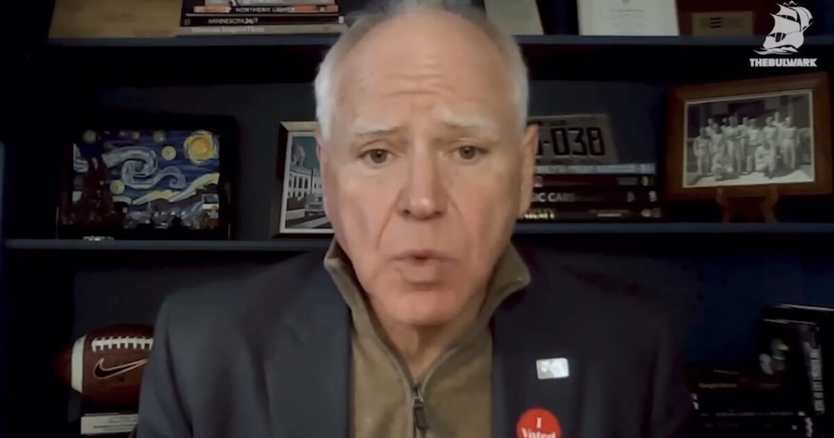 Man wearing a jacket and "I Voted" sticker speaks in front of a bookshelf with various items and artwork.