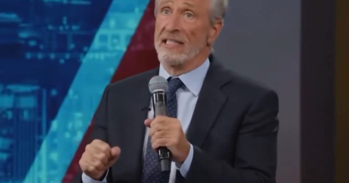 Comedian delivering a stand-up performance on stage, engaging the audience with humor and commentary, wearing a suit and holding a microphone.