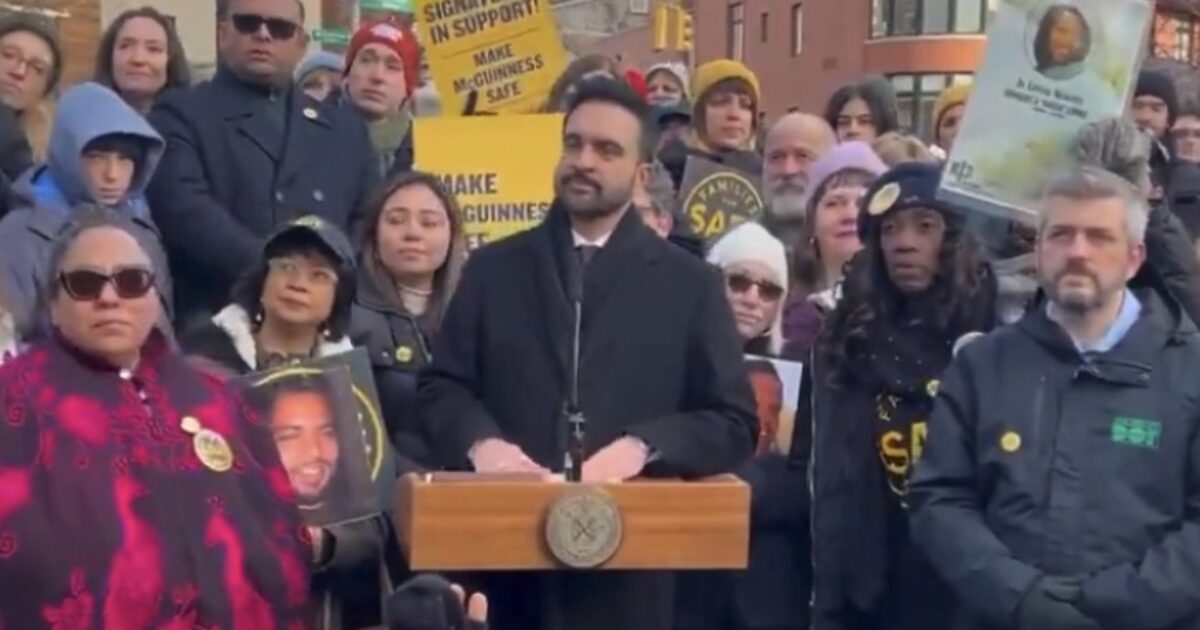 A diverse group of supporters gathers at a rally, with a speaker at a podium addressing the crowd, holding signs advocating for safety and justice.