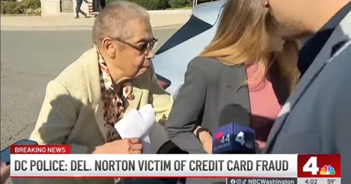 Elderly woman speaking to reporters about being a victim of credit card fraud, with police presence and media coverage in the background.