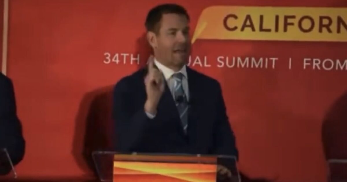 Speaker at the California 34th Annual Summit presents key points, gesturing with one hand, against a vibrant red backdrop.