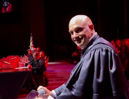 Smiling speaker in graduation attire addresses an audience at a ceremony, with a vibrant backdrop and flags visible.
