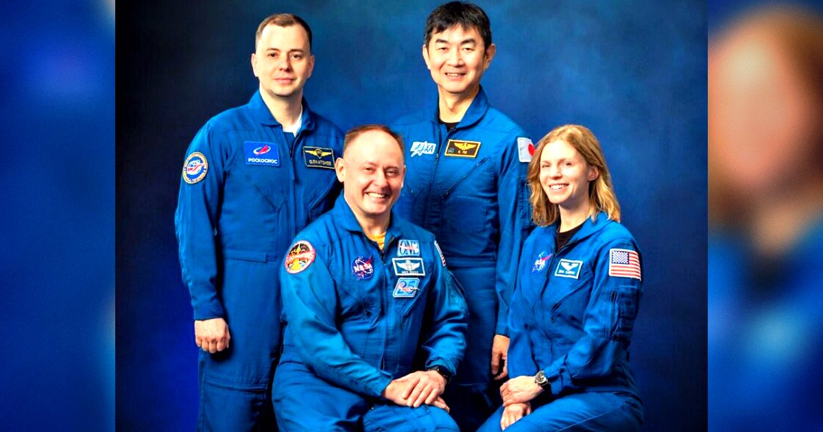 Group portrait of four astronauts in blue flight suits, smiling against a blue background, representing international collaboration in space exploration.