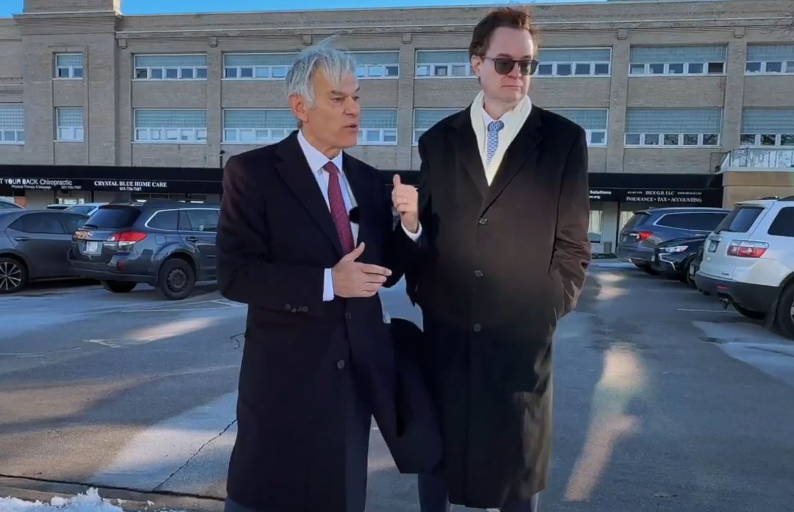 Two men in formal attire stand outdoors, discussing in front of a parking lot with various vehicles and a building in the background.