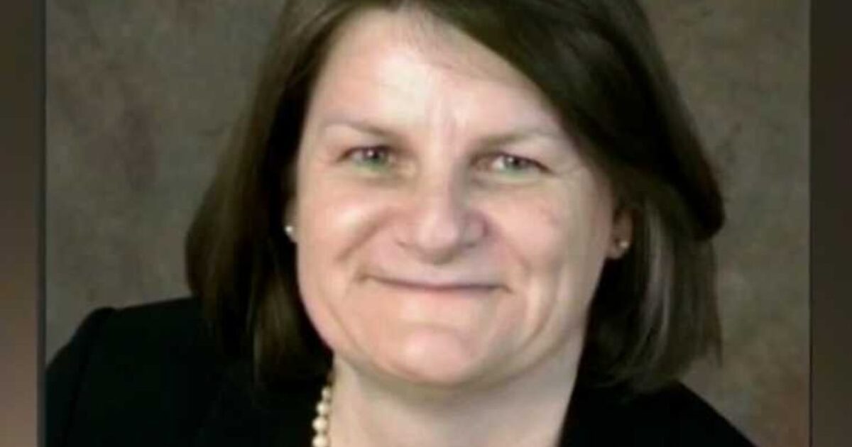 Smiling woman with shoulder-length brown hair wearing a black blazer and pearl necklace, set against a neutral background.