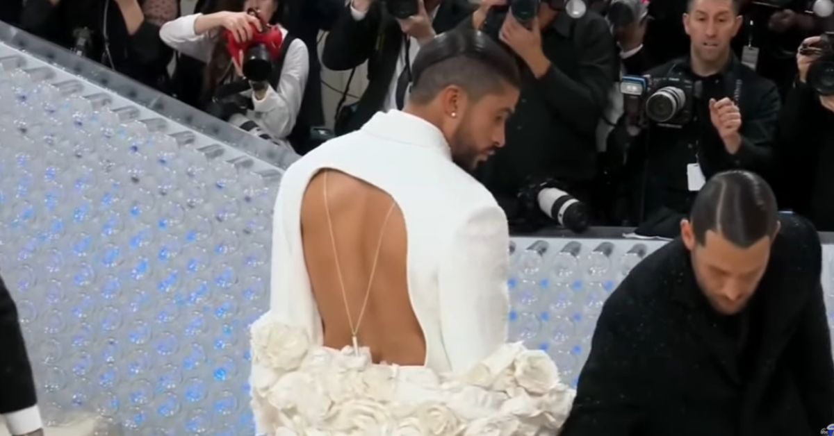 A man in a stylish white suit with a backless design adorned with roses walks the runway, surrounded by photographers capturing the moment.
