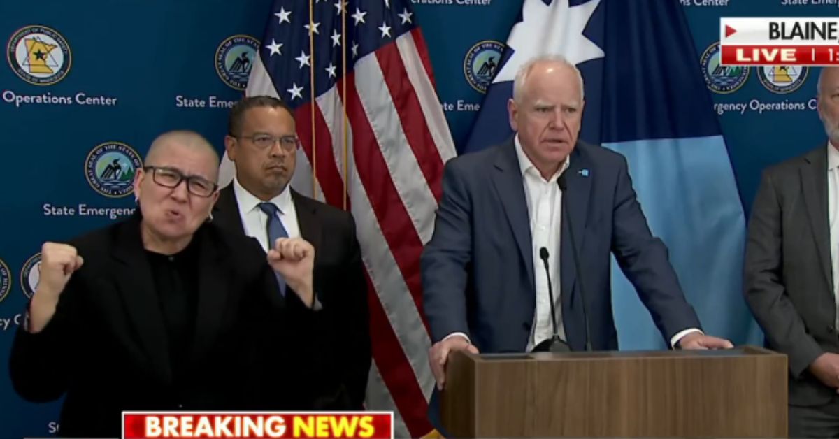 Press conference featuring state officials discussing emergency operations, with an interpreter providing sign language support, set against a backdrop of flags and emergency center logos.