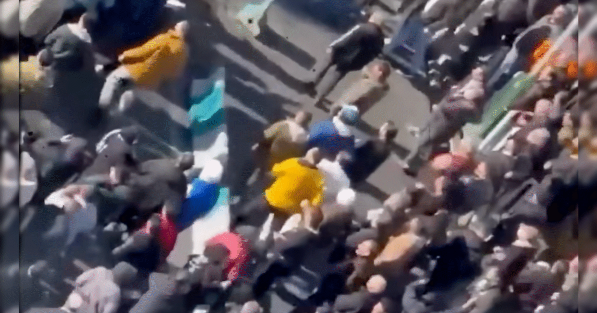 Crowd of people gathered in a public space, some wearing bright colored clothing, during a daytime event or protest.