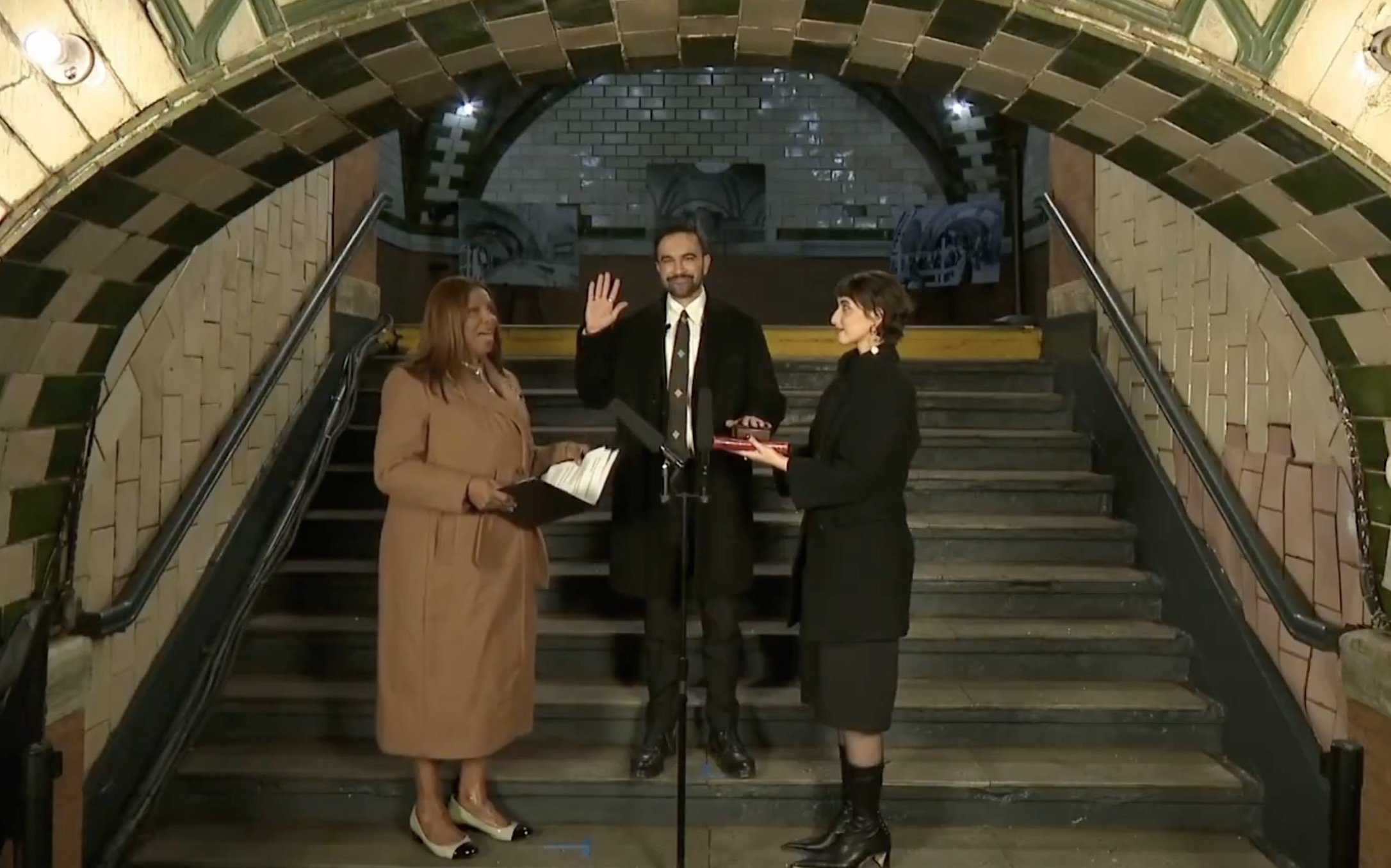 Ceremony in a subway station featuring a man taking an oath, flanked by two women holding documents, with an ornate archway in the background.