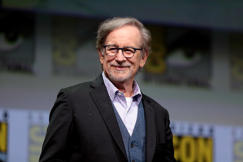 Filmmaker Stephen Spielberg smiling on stage at a major event, wearing glasses and a black suit, with a colorful background featuring event logos.