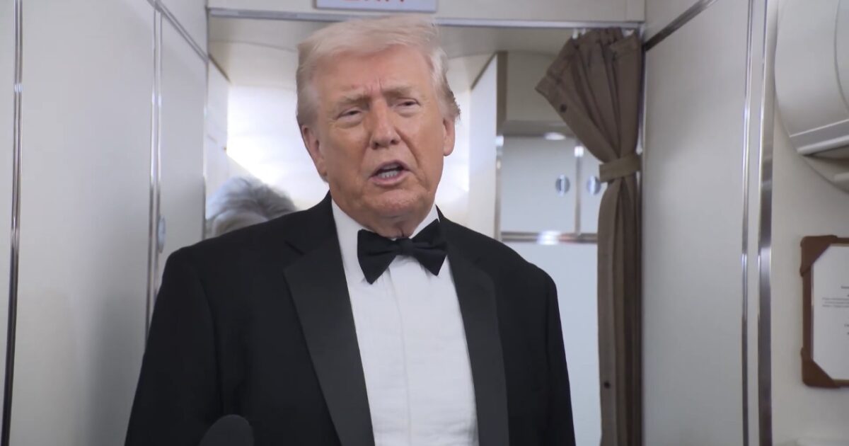 Donald Trump speaking in a tuxedo inside an aircraft, with an exit sign visible in the background.