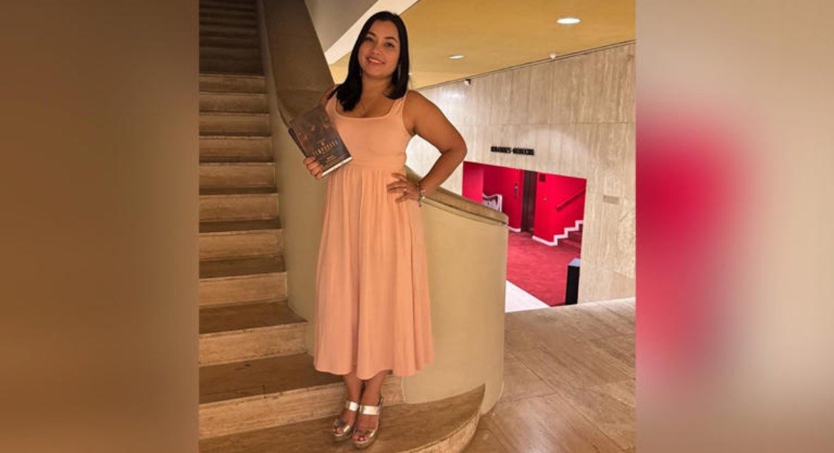 Woman in a pink dress holding a book, standing on a staircase in a stylish interior with red accents.