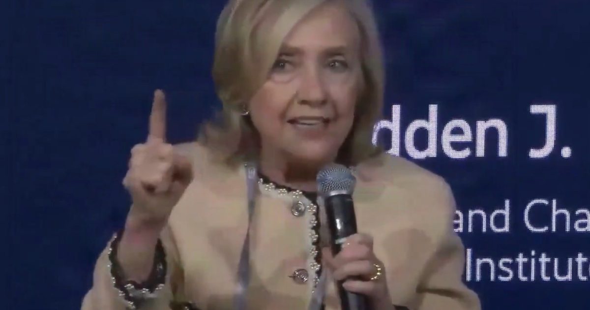 Hillary Clinton speaking passionately at a conference, holding a microphone and gesturing with one hand to emphasize her point.