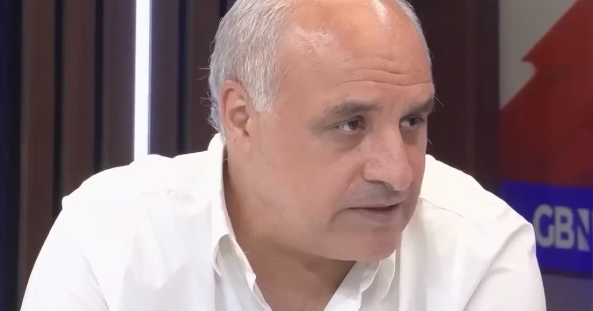 A man in a white shirt is engaged in a discussion, sitting at a table with modern studio lighting and a backdrop featuring a logo.