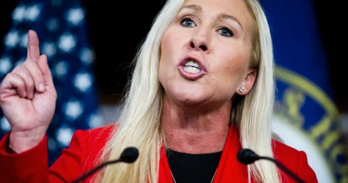 Marjorie Taylor Greene passionately speaks at a press conference, gesturing with her hand and wearing a red blazer against a backdrop of American flags.