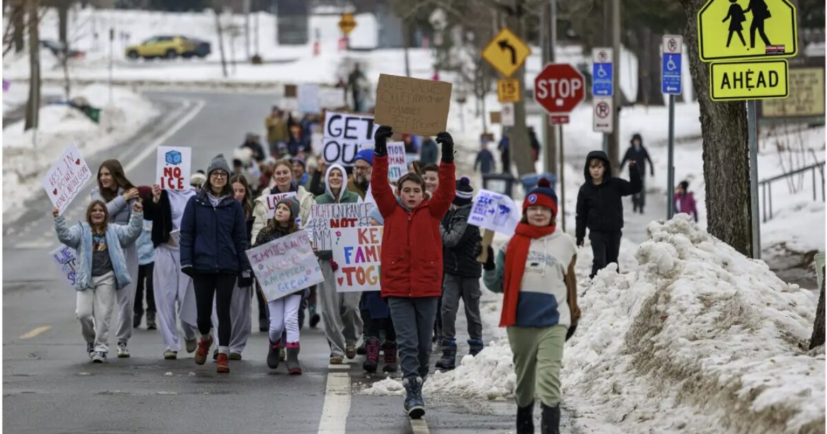 Students and community members march in winter attire holding signs advocating for social change on a snowy street.