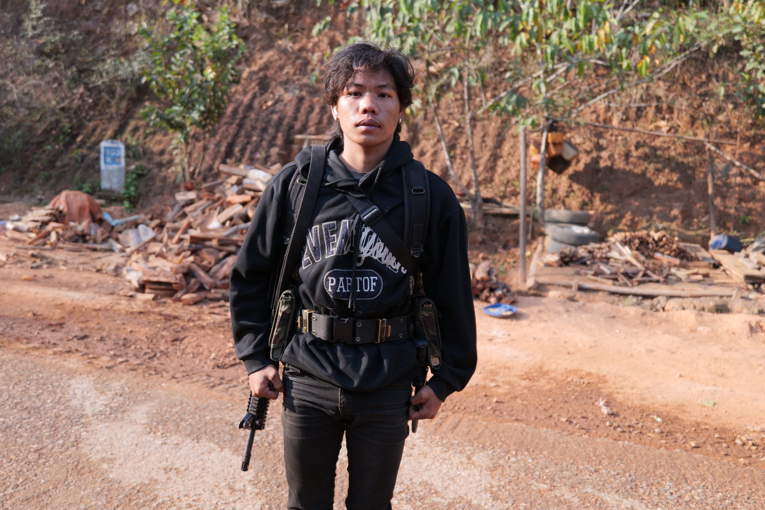 Young man dressed in tactical gear standing on a dirt road with a wooded background, holding a firearm and wearing wireless earbuds.